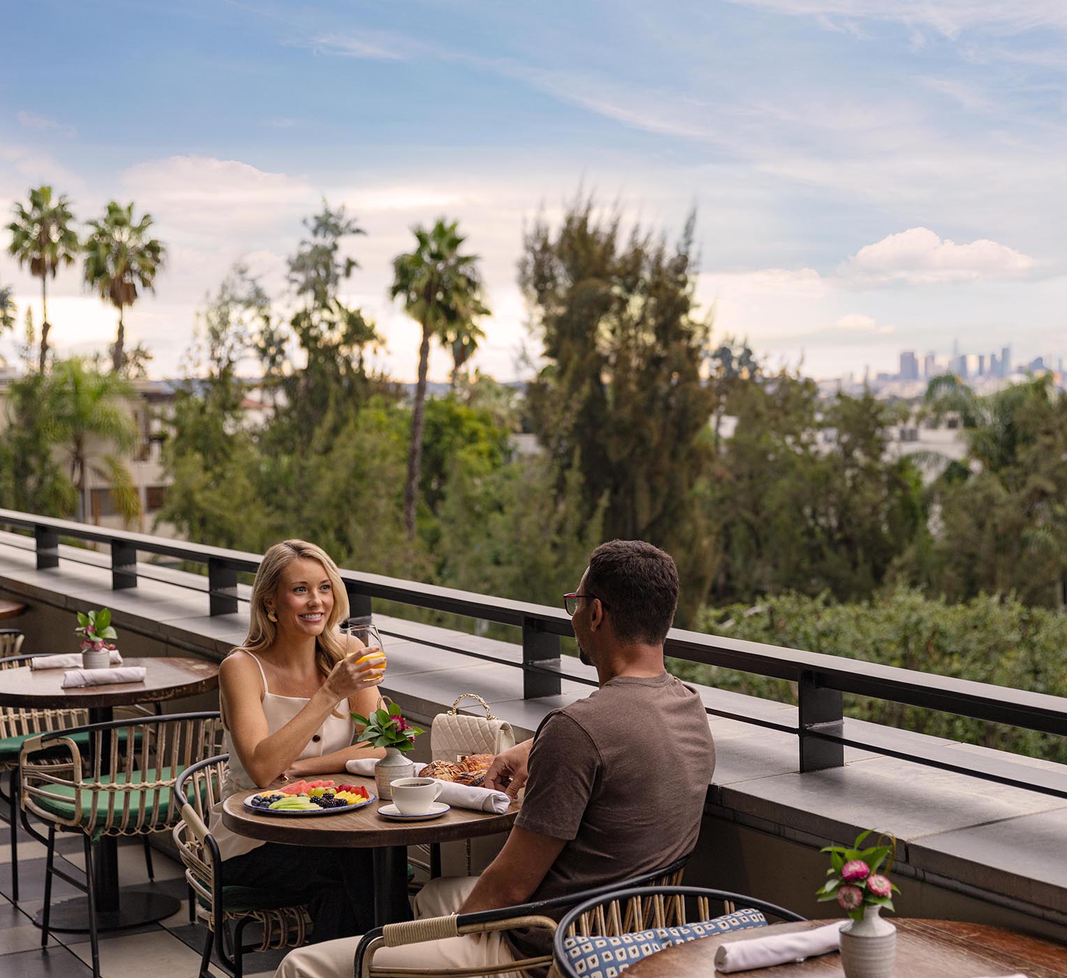 Couple dining on an outdoor terrace at The Sun Rose hotel, surrounded by lush greenery with sweeping Los Angeles skyline views in the distance.