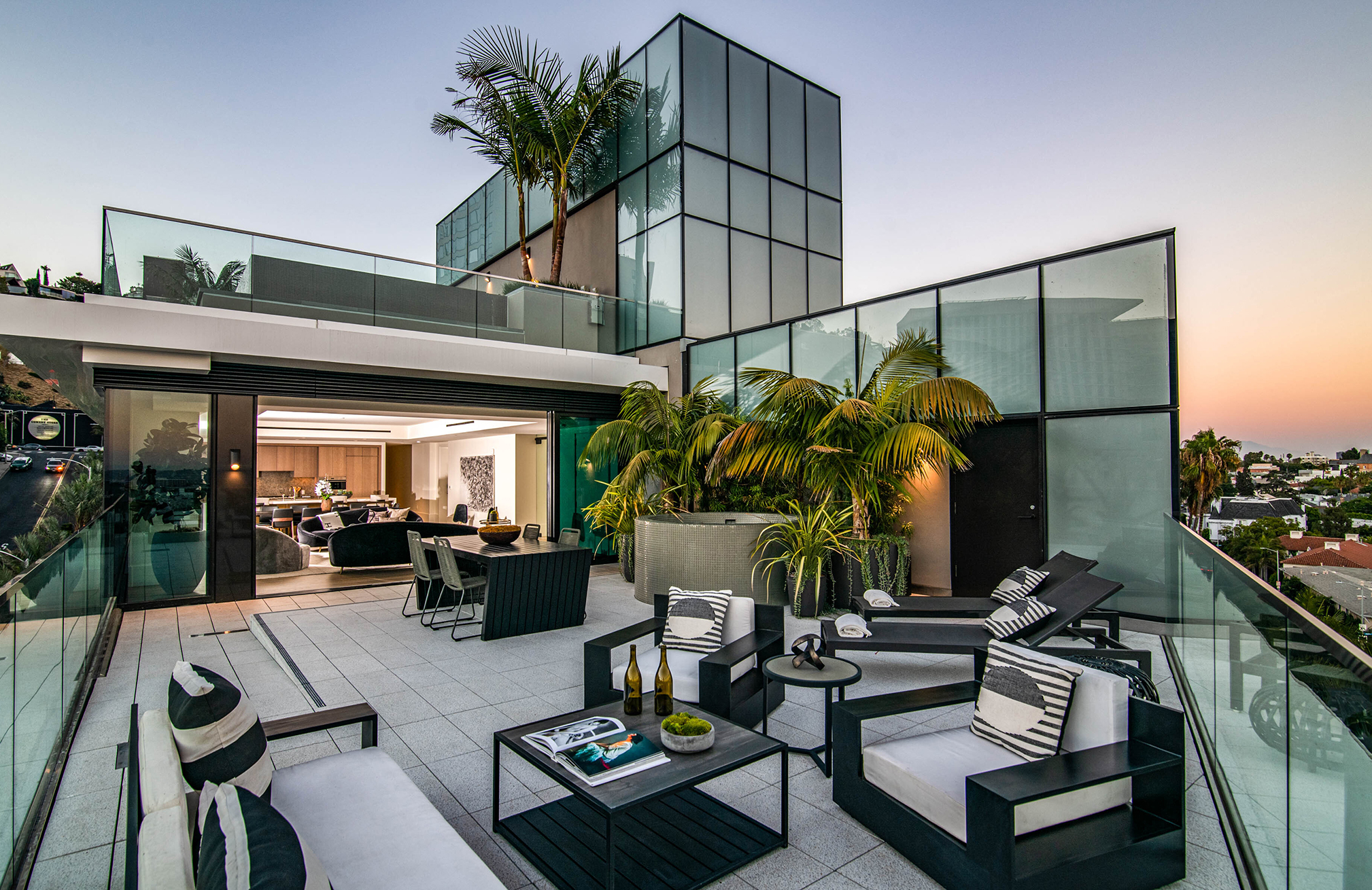 Modern rooftop terrace of a luxury residence featuring glass railings, outdoor lounge seating, and a dining area, with sliding doors opening to a bright, contemporary interior. Lush potted palms and landscaping add privacy, while the elevated setting offers views over a residential neighborhood at sunset.